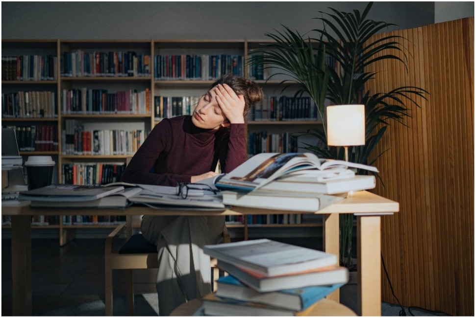 Stressed college student surrounded by books in a library while choosing a research paper topic.